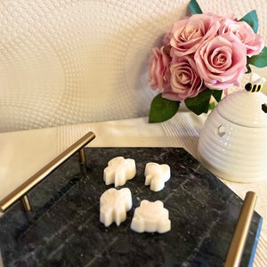 May include: A black marble tray with a gold handle, holding four white wax melts shaped like leaves. The tray is on a white surface with a white ceramic beehive-shaped wax warmer and a bouquet of pink roses.