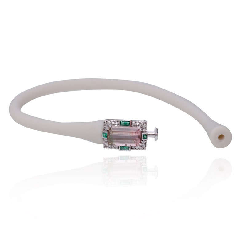 May include: A white rubber bracelet with a rectangular silver clasp featuring a pink and green gemstone surrounded by smaller diamonds and emeralds.