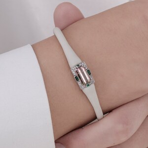 May include: A white silicone bracelet featuring a pink and green gemstone set in a silver frame. The gemstone is surrounded by smaller diamonds and green stones.