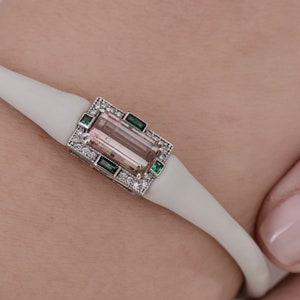 May include: A white bracelet featuring a pink and brown gemstone set in a silver frame with small green and white stones.