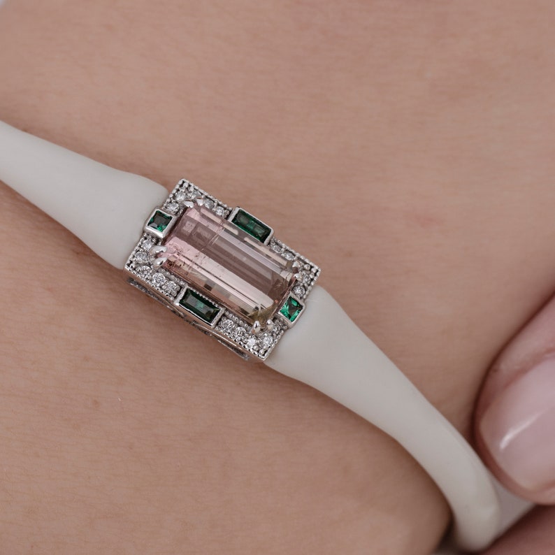 May include: A white bracelet featuring a pink and brown gemstone set in a silver frame with small green and white stones.