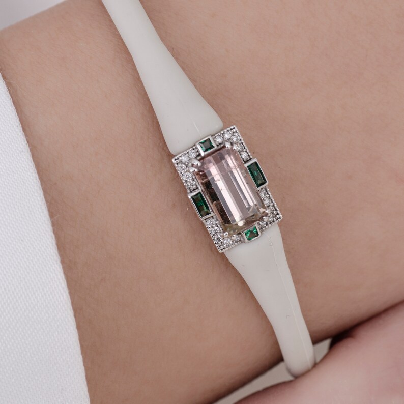 May include: A white bracelet featuring a rectangular pink and white gemstone set in a silver frame with green stones. The gemstone is surrounded by smaller white stones.