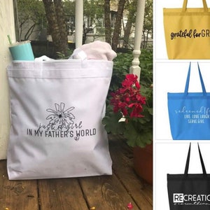 May include: Four tote bags with different designs and colors. The first bag is white with black text that says "just a girl in my father's world". The second bag is yellow with black text that says "grateful for GRACE". The third bag is blue with white text that says "redeemed free live love laugh serve give". The fourth bag is black with white text that says "RECREATION 2 Corinthians 5:17".