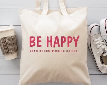 May include: A white canvas tote bag with the text "BE HAPPY" in pink. The text below reads "READ BOOKS ♥ DRINK COFFEE". The bag is sitting on a white wooden surface with a white coffee cup and two white sneakers.