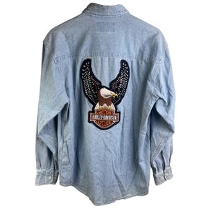 May include: Light blue denim shirt with long sleeves. Features a Harley-Davidson patch on the back with an eagle design and the brand's logo. The shirt has a button-down front and cuffs.