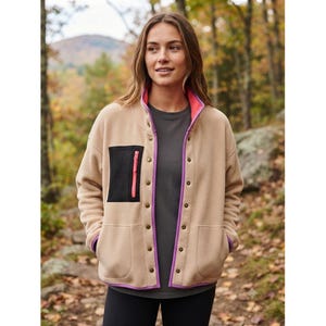 May include: A beige fleece jacket featuring a snap-button front closure, a black mesh chest pocket with a bright pink zipper, and purple trim along the collar, placket, and hem. The jacket is worn over a dark gray shirt and black pants.