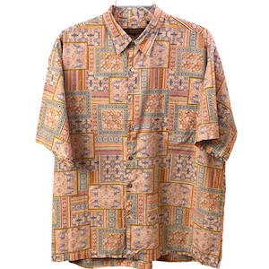 May include: Short-sleeved button-down shirt with a patchwork design. The shirt features a repeating pattern of squares and rectangles in shades of peach, brown, blue, and gold. The shirt is hanging on a white background.