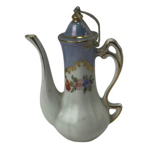 May include: A ceramic coffee pot ornament with a light blue lid and handle, and a white body. The ornament is decorated with gold trim and a floral design in red, pink, and blue. A gold string is attached for hanging.
