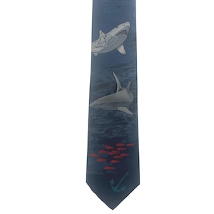 May include: A dark blue necktie featuring a printed design of two grey sharks swimming in deep water, a small school of tiny red fish, and a single teal anchor icon positioned near the pointed bottom edge of the fabric accessory.