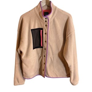 May include: A tan fleece jacket with a high collar and snap closures. The jacket features a black mesh pocket with a pink zipper, purple trim, and a red interior lining. The jacket is hanging on a wooden hanger.