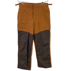 May include: Brown work pants with a contrasting dark brown lower leg panel. The pants have two front pockets and two back pockets. They are hanging on a white hanger.