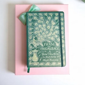 May include: A green hardcover journal with a peacock design and the title "Pride and Prejudice" by Jane Austen. The journal is on a pink gift box with a green ribbon.