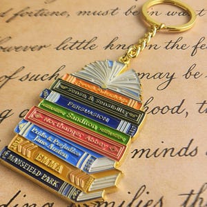 May include: A gold-colored keychain featuring a stack of colorful books with the titles "Sense & Sensibility", "Persuasion", "Sanditon", "Northanger Abbey", "Pride & Prejudice", "Emma", and "Mansfield Park".