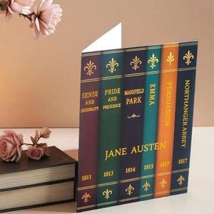 May include: A greeting card designed to look like a stack of classic books by Jane Austen. The card features titles like "Sense and Sensibility" and "Pride and Prejudice" in gold lettering on colorful spines. Decorative floral accents are visible.