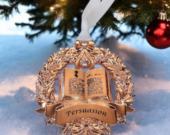 May include: A gold-colored Christmas ornament featuring an open book design with the word "Persuasion" on a banner. The ornament is surrounded by a decorative wreath and has a white ribbon for hanging. A red ornament and Christmas tree are in the background.