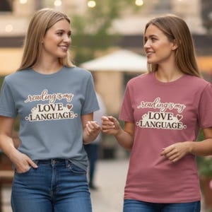 Può includere: Due donne indossano magliette con la scritta "reading is my LOVE LANGUAGE". Una maglietta è blu polveroso, l'altra è malva. Entrambe le magliette hanno lo stesso design in corsivo con un cuore e un piccolo motivo floreale.