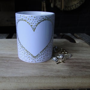 May include: White ceramic mug with a gold heart design. The mug is sitting on a wooden surface.
