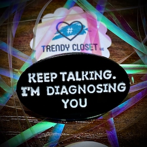 May include: Black enamel pin with white text that reads "Keep Talking. I'm Diagnosing You".