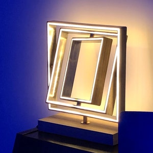 Può includere: Una lampada da tavolo moderna e minimalista con una cornice dorata e una base bianca. La lampada presenta una serie di quadrati sovrapposti con luci LED che creano una luce calda e ambientale.