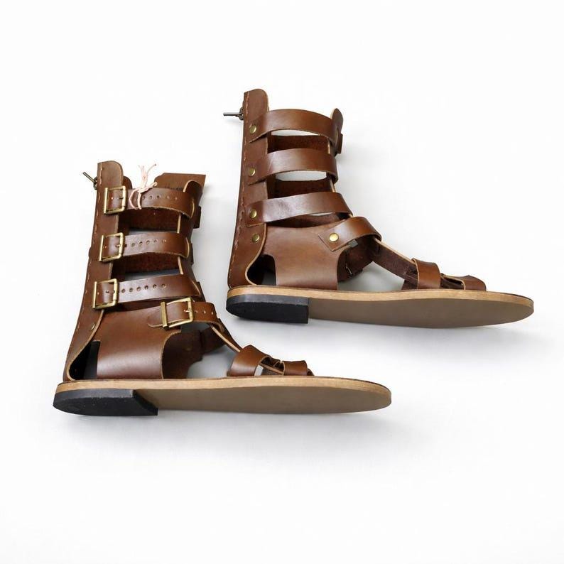 May include: A pair of brown leather gladiator sandals featuring a high-cut design with multiple horizontal straps, brass-toned metal buckles, and a flat sole. The sandals have an open-toe construction and a secure back panel with visible stitching.