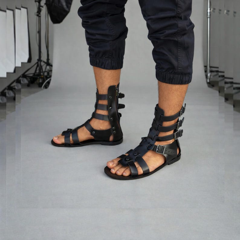 May include: Black leather gladiator sandals featuring a high-cut design with multiple horizontal straps, silver-toned metal buckles, and a back zipper closure. The sandals have a flat sole and an open-toe construction, extending up to the lower calf.