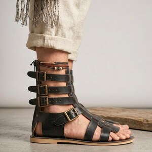 May include: A black leather gladiator sandal featuring a high-cut design with multiple straps, antique brass-toned metal buckles, a back zipper closure, and a flat tan sole. The sandal extends up the ankle with adjustable straps for a secure fit.