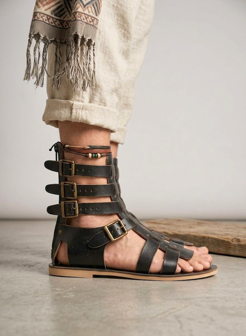 May include: A black leather gladiator sandal featuring a high-cut design with multiple straps, antique brass-toned metal buckles, a back zipper closure, and a flat tan sole. The sandal extends up the ankle with adjustable straps for a secure fit.