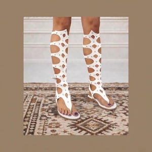 May include: White gladiator sandals with knee-high cut-out design. The sandals feature a thong style toe strap and are adorned with small silver studs. The sandals are worn on a patterned rug.