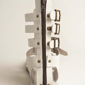 May include: A white gladiator sandal featuring a vertical brown zipper closure along the back heel panel. The sandal includes three horizontal straps secured with bronze-toned metal buckles on the side and is accented with small bronze rivets.