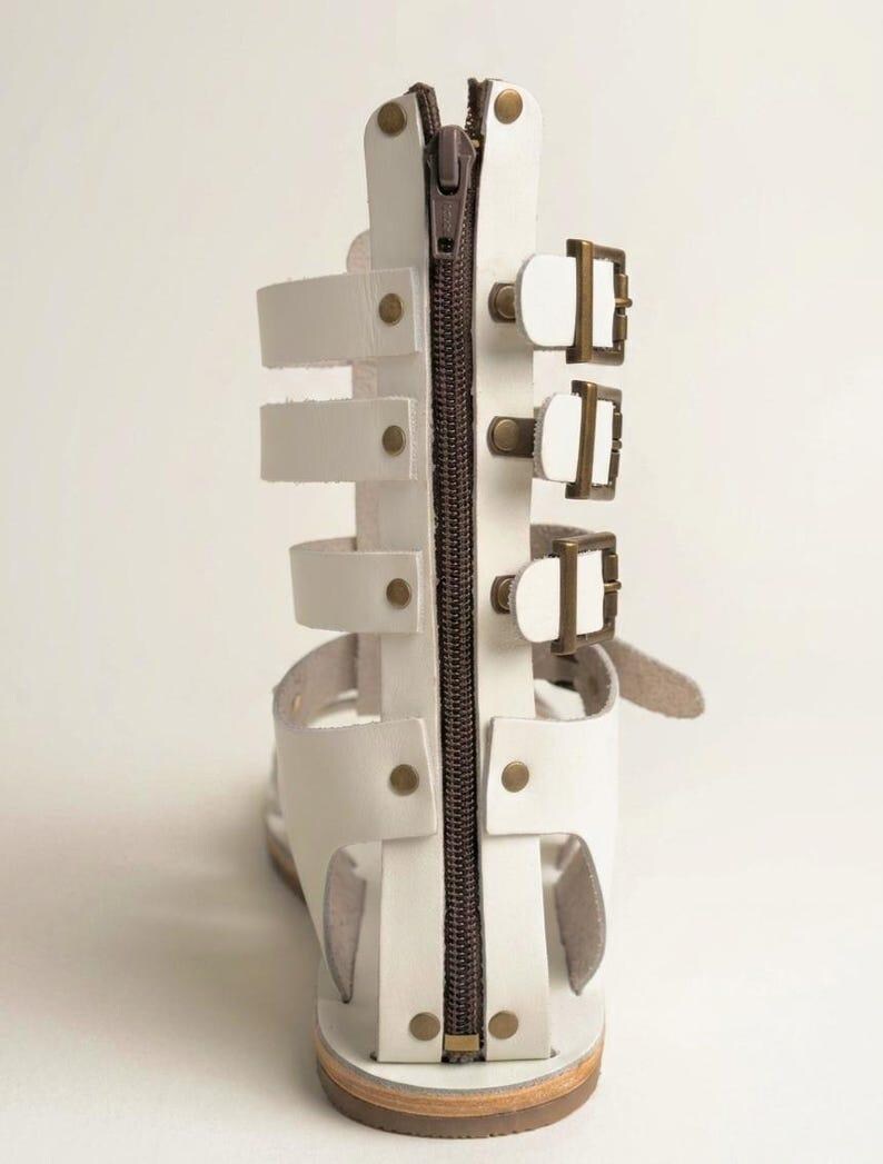May include: A white gladiator sandal featuring a vertical brown zipper closure along the back heel panel. The sandal includes three horizontal straps secured with bronze-toned metal buckles on the side and is accented with small bronze rivets.