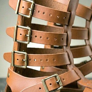 May include: A pair of tan leather gladiator sandals featuring a high-cut design with multiple horizontal straps. Each strap is secured with a metallic buckle, providing an adjustable fit. The construction includes a flat sole and open sides.
