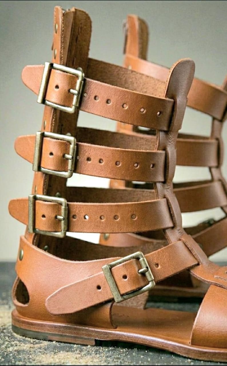May include: A pair of tan leather gladiator sandals featuring a high-cut design with multiple horizontal straps. Each strap is secured with a metallic buckle, providing an adjustable fit. The construction includes a flat sole and open sides.