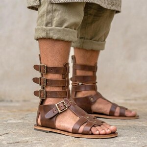 May include: Brown leather gladiator sandals featuring a high-cut design with multiple horizontal straps, antique brass-toned metal buckles, and a flat sole. The footwear extends above the ankle and secures with a series of adjustable side buckles.