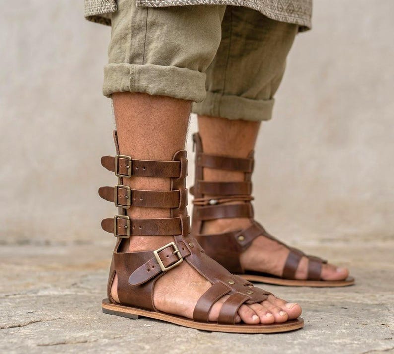 May include: Brown leather gladiator sandals featuring a high-cut design with multiple horizontal straps, antique brass-toned metal buckles, and a flat sole. The footwear extends above the ankle and secures with a series of adjustable side buckles.