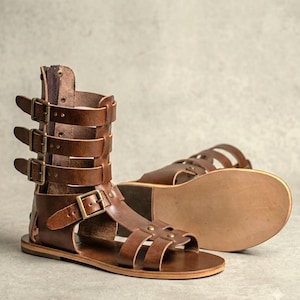 May include: A pair of brown leather gladiator sandals featuring a high-cut shaft with three adjustable buckle straps, a zipper closure at the back, and a flat sole. The design includes additional straps across the foot secured with metal buckles.
