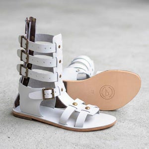 May include: A pair of white gladiator sandals featuring a high-cut design with multiple horizontal straps, bronze-toned metal buckles, and a rear zipper closure. The flat sole displays a circular stamp with the text HANDMADE and the size 39.