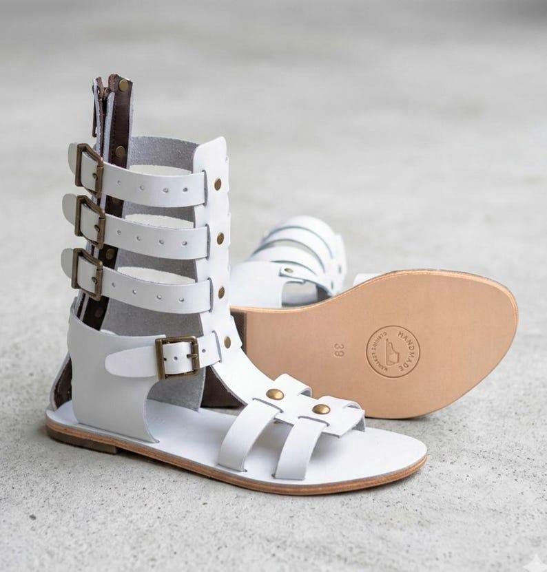 May include: A pair of white gladiator sandals featuring a high-cut design with multiple horizontal straps, bronze-toned metal buckles, and a rear zipper closure. The flat sole displays a circular stamp with the text HANDMADE and the size 39.