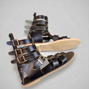 May include: A pair of black gladiator sandals featuring multiple horizontal straps with antique brass-toned metal buckles. The sandals have a high-cut ankle design with a vertical back zipper closure and a flat, light-colored sole for a secure fit.