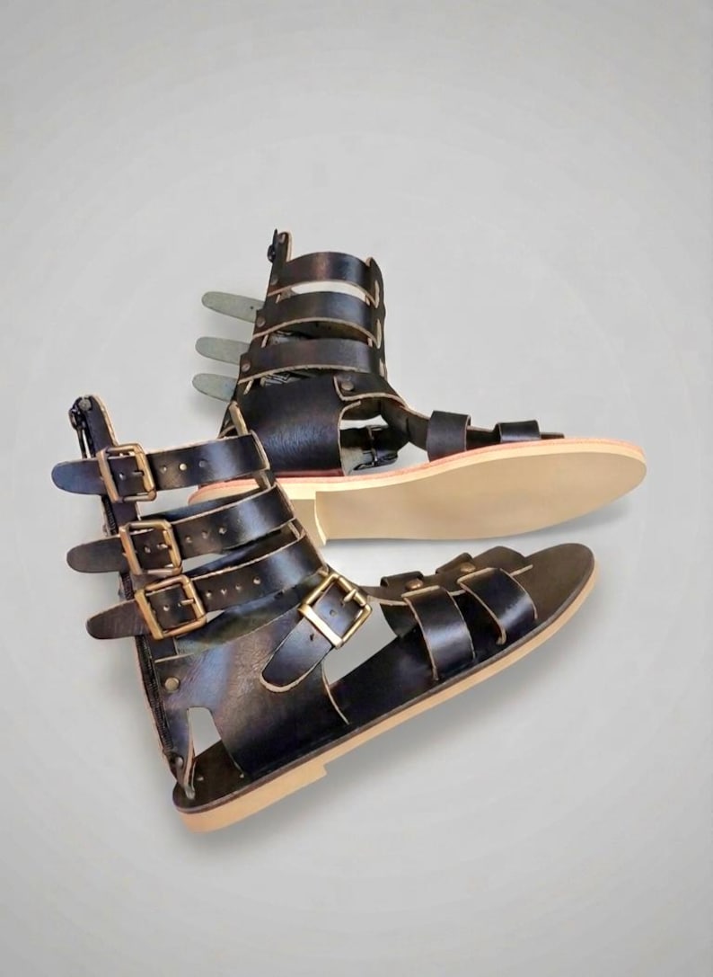 May include: A pair of black gladiator sandals featuring multiple horizontal straps with antique brass-toned metal buckles. The sandals have a high-cut ankle design with a vertical back zipper closure and a flat, light-colored sole for a secure fit.