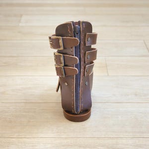 May include: A dark brown leather boot featuring a vertical center zipper and three horizontal buckled straps on each side. The boot has visible stitching along the edges and metallic rivets securing the straps to the main structure of the footwear.