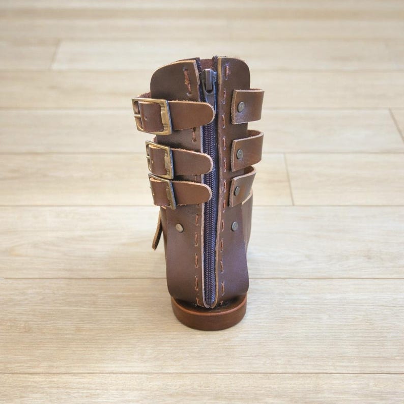 May include: A dark brown leather boot featuring a vertical center zipper and three horizontal buckled straps on each side. The boot has visible stitching along the edges and metallic rivets securing the straps to the main structure of the footwear.