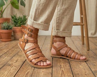 May include: Tan leather gladiator sandals featuring multiple horizontal straps across the foot and three adjustable ankle straps with metal buckles. The footwear has a flat sole and an open-toe design, providing a secure fit around the lower leg.