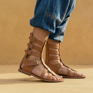 May include: A pair of tan leather gladiator sandals featuring multiple horizontal straps that extend up the ankle, each secured with a metal buckle. The sandals have an open-toe design and a flat sole, worn with loose-fitting blue denim trousers.
