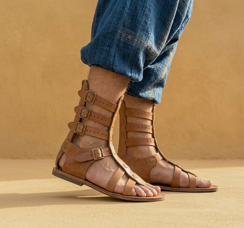 May include: A pair of tan leather gladiator sandals featuring multiple horizontal straps that extend up the ankle, each secured with a metal buckle. The sandals have an open-toe design and a flat sole, worn with loose-fitting blue denim trousers.