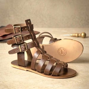 Könnte beinhalten: Ein Paar braune Ledersandalen im Gladiatorenstil mit drei verstellbaren Knöchelriemen und messingfarbenen Metallschnallen. Die flache Sohle ist hellbraun und im Fersenbereich mit einem kreisförmigen Stempelaufdruck versehen.