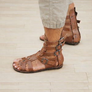 May include: Brown leather gladiator sandals featuring multiple horizontal straps across the foot and ankle, secured with silver-toned metal buckles. The design includes a high-cut back panel with vertical straps and visible metal rivets along the sides.