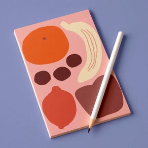 Può includere: Un blocco note rosa con illustrazioni di frutta colorate, tra cui un'arancia, una banana e un limone, accanto a una matita bianca. I disegni della frutta sono nei toni dell'arancione, marrone e beige. Il blocco note è su una superficie blu.
