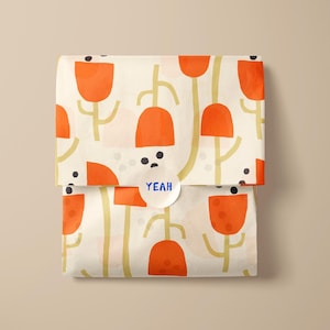 Könnte beinhalten: Ein gefaltetes Paket, verpackt in cremefarbenem Papier mit einem sich wiederholenden Muster aus orangefarbenen tulpenartigen Formen und goldenen Stielen. Ein weißer runder Aufkleber mit dem Wort "YEAH" in Blau ist vorne angebracht.