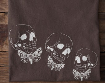 Puede incluir: Una camiseta marrón con un gráfico blanco de tres calaveras con polillas sobre un fondo de madera marrón.