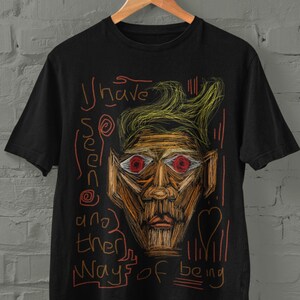 Puede incluir: Camiseta negra con un gráfico de una cara estilizada con ojos rojos y el texto "He visto tantas formas de ser".